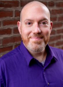 A bearded man with a shaed head wears a purple shirt while smiling in front of a brick wall.